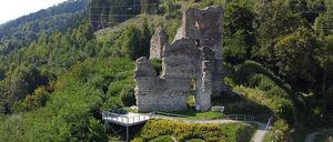 Auf einem Hügel steht eine alte Burgruine, umgeben von üppiger Grünfläche. Ein herzförmiger Garten aus grünen und gelben Pflanzen fügt einen Hauch von Liebe hinzu. Die Burg, teilweise restauriert, hat eine Steinstruktur mit einer nahegelegenen Holzplattform.