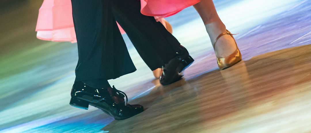 Ein Paar tanzt auf einer Bühne, die Frau in einem rosa Kleid und der Mann in Schwarz. Das Kleid der Frau weht im Wind, und sie sind in einer Bewegung.