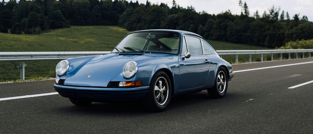 Ein blauer Porsche 911 fährt auf einer Autobahn mit einer malerischen Aussicht auf einen begrünten Hügel und Bäume unter einem bewölkten Himmel.