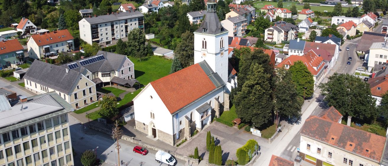 Luftaufnahme einer kleinen Stadt mit einer Kirche in der Mitte, umgeben von Wohngebäuden, grünen Hügeln und einem klaren blauen Himmel. Die Kirche hat ein rotes Dach und einen hohen Turm.