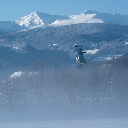 Eine neblige Landschaft mit einem verschneiten Bergrücken und einem Turm in der Mitte, umgeben von Bäumen.