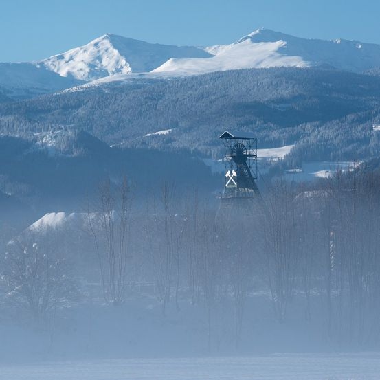 Eine neblige Landschaft mit einem verschneiten Bergrücken und einem Turm in der Mitte, umgeben von Bäumen.