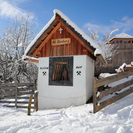 Eine kleine Kapelle namens St. Barbara, bedeckt mit Schnee. Ein hölzerne Kreuz auf dem Dach und ein kleines Fenster mit einem Metallgitter. Umgeben von Holzzäunen.