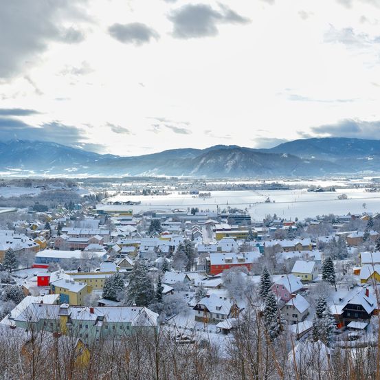 Eine verschneite Stadt mit Häusern und Bergen in der Ferne. Der Himmel ist bewölkt.