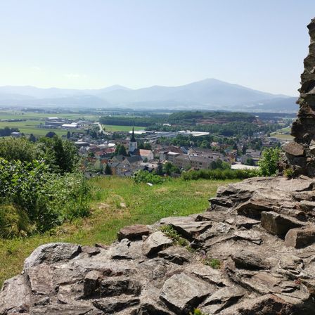 Luftaufnahme einer kleinen Stadt mit einem Kirchturm. Grüne Felder, Bäume und entfernte Berge. Heller Himmel.