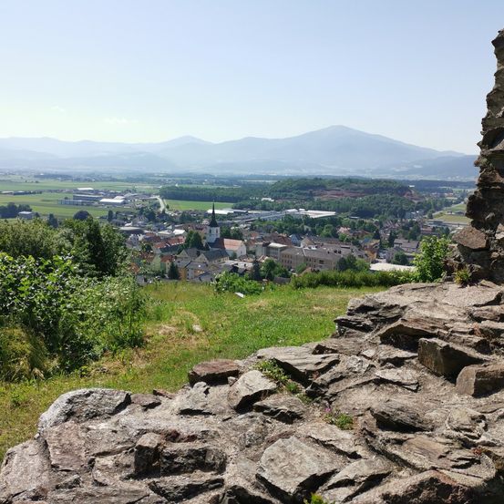 Luftaufnahme einer kleinen Stadt mit einem Kirchturm. Grüne Felder, Bäume und entfernte Berge. Heller Himmel.