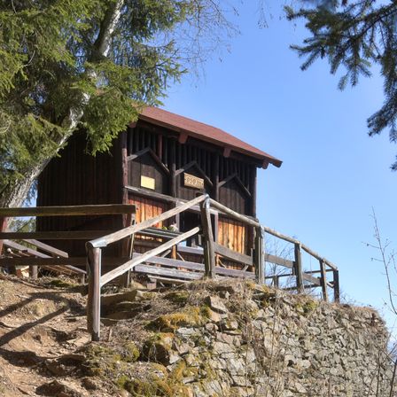 Ein kleines Holzgebäude mit rotem Dach steht auf einem felsigen Hügel, umgeben von Bäumen und einem Holzgeländer. Das Gebäude hat ein Schild mit der Nummer 899.