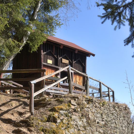 Ein kleines Holzgebäude mit rotem Dach steht auf einem felsigen Hügel, umgeben von Bäumen und einem Holzgeländer. Das Gebäude hat ein Schild mit der Nummer 899.