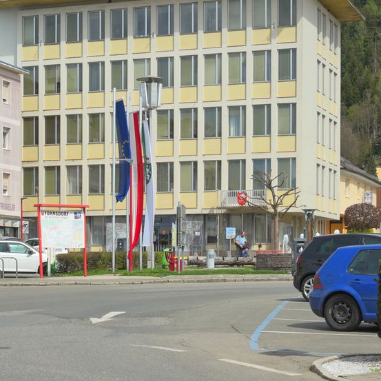 Ein großes mehrstöckiges Gebäude mit gelben Akzenten steht vor einer Straße mit geparkten Autos und Flaggen.