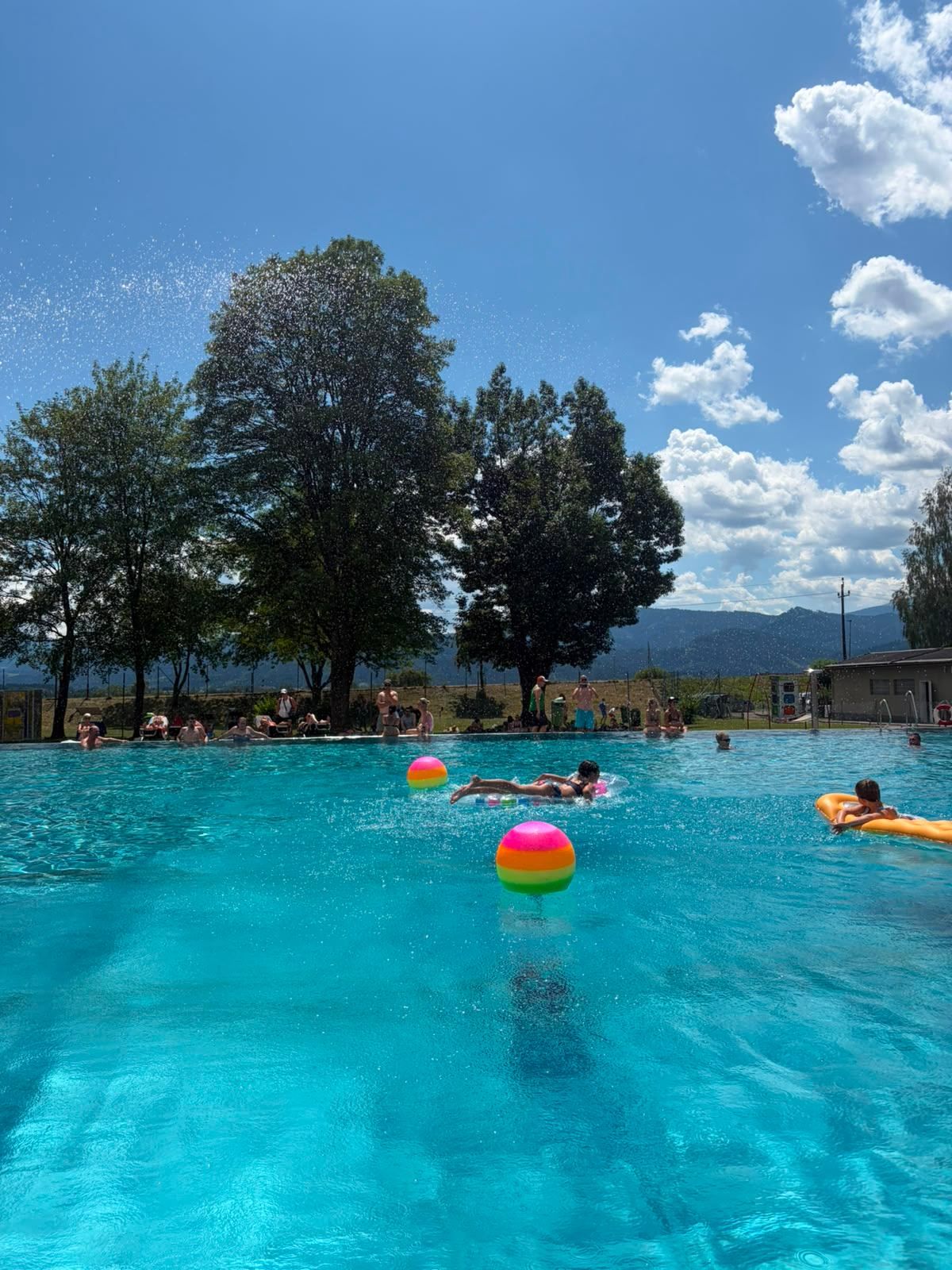 A large outdoor pool with people swimming and floating on inflatable toys under a blue sky with clouds and trees in the background.