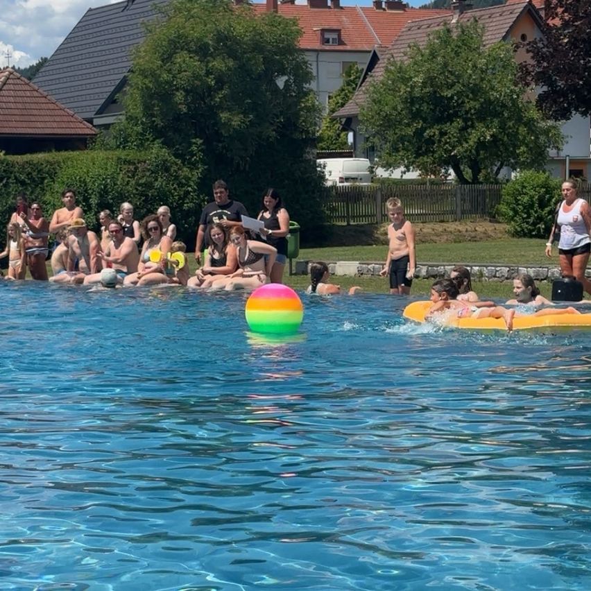 A group of people are gathered around a pool, some sitting on the edge and others floating on inflatable rafts. A vibrant multicolored ball floats in the water.