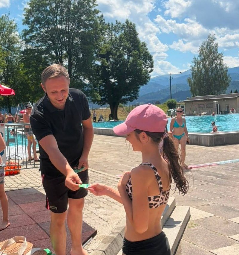 A man hands a green ticket to a girl in a pink hat at an outdoor pool. In the background, people swim in the pool, and mountains are visible.