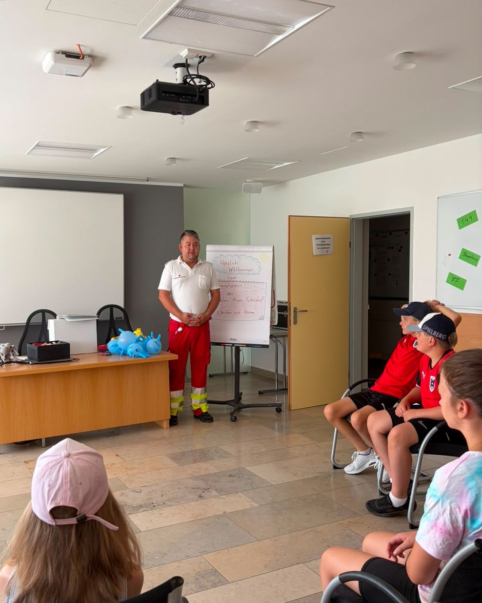 A man in a white shirt and red pants stands in front of a whiteboard, addressing a group of children sitting in chairs. The room has a projector, a desk, and a whiteboard with writing on it.