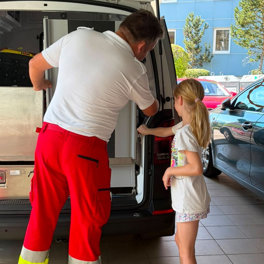 A man in red pants and a white shirt is opening the back door of a van while a young girl watches.