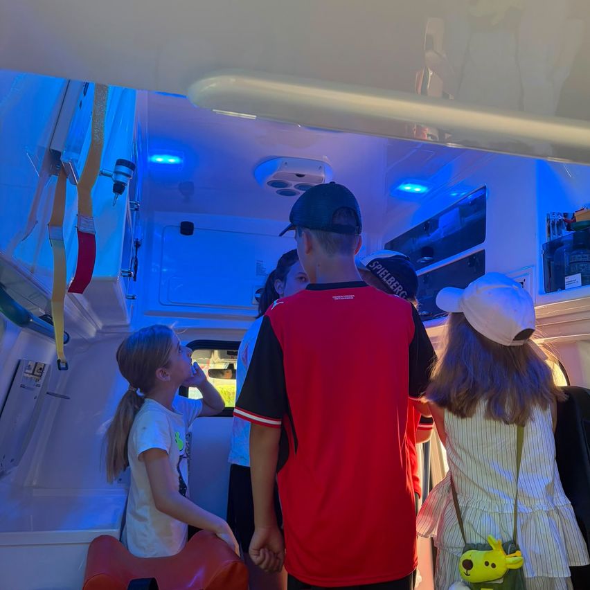 Inside an ambulance, a group of children and adults are looking around. The interior is lit with blue lights, and medical equipment is visible.