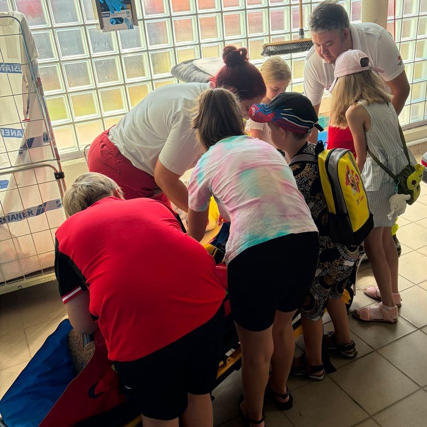 A group of children and adults are gathered around a stretcher, possibly in a hospital setting, with one adult in a red shirt bending over the stretcher.