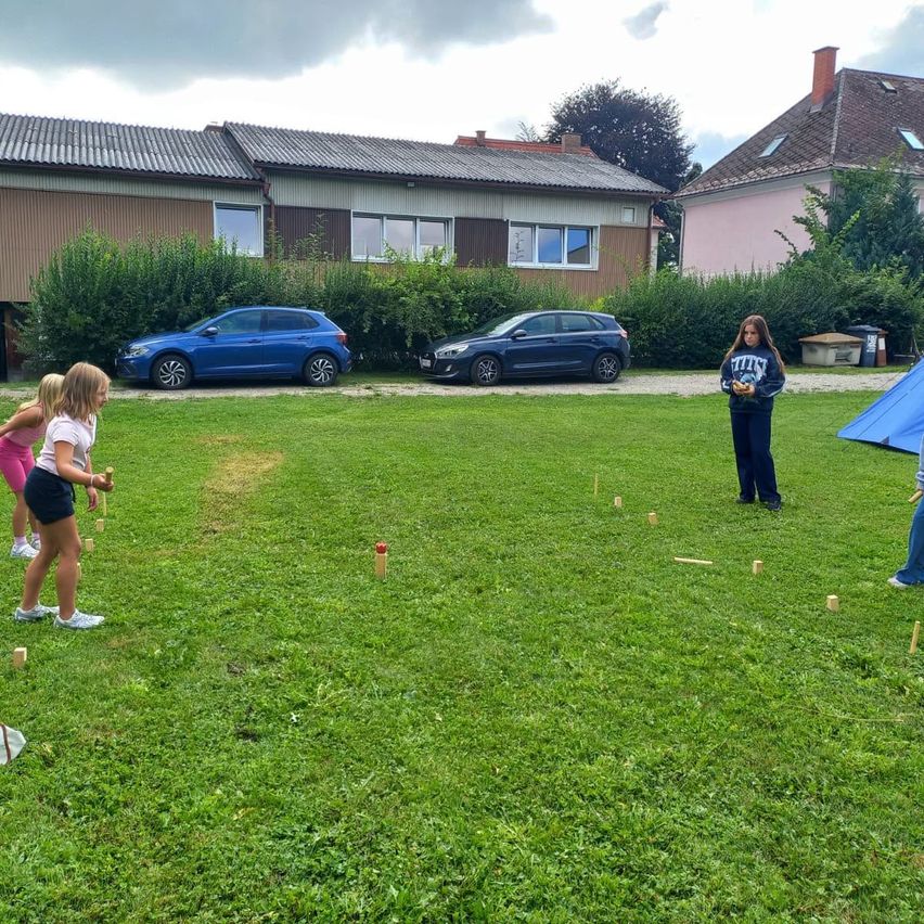 Drei Mädchen spielen ein Kegelspiel auf einem Rasenfeld, mit Häusern und Autos im Hintergrund.