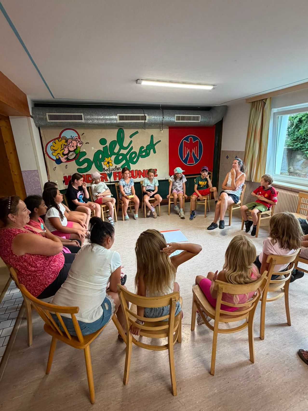 Eine Gruppe von Kindern und Erwachsenen sitzt in einem Kreis in einem Raum mit einem Banner, das 'Spiel-Gesund' und 'Wilder FFC' besagt. Sie hören einer Frau zu, die spricht. Der Raum hat Holzstühle, ein Fenster und einen Heizkörper.