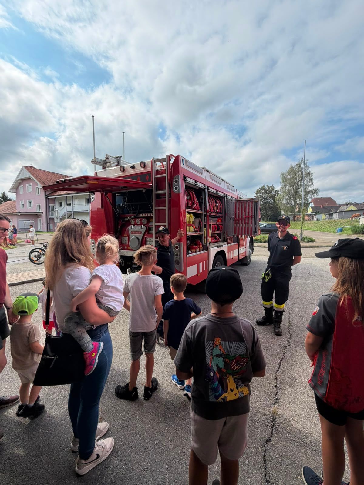Eine Gruppe von Kindern und Erwachsenen versammelt sich um einen roten Feuerwehrwagen, wobei zwei Feuerwehrleute ihnen etwas erklären. Die Szene spielt im Freien bei bewölktem Himmel mit Häusern im Hintergrund.