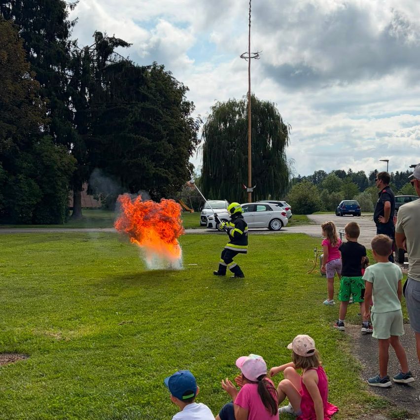 Ein Feuerwehrmann löscht ein Feuer auf einer Wiese, während Kinder und Erwachsene zuschauen.
