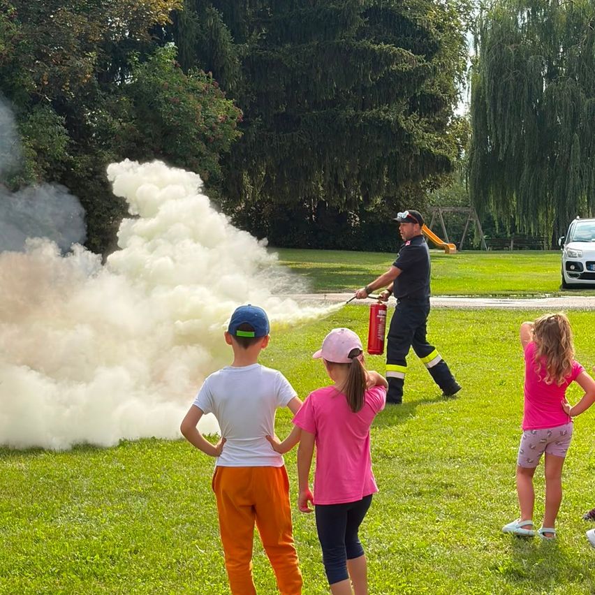 Ein Feuerwehrmann sprüht Schaum aus einem Feuerlöscher, während Kinder in einem Park zusehen.