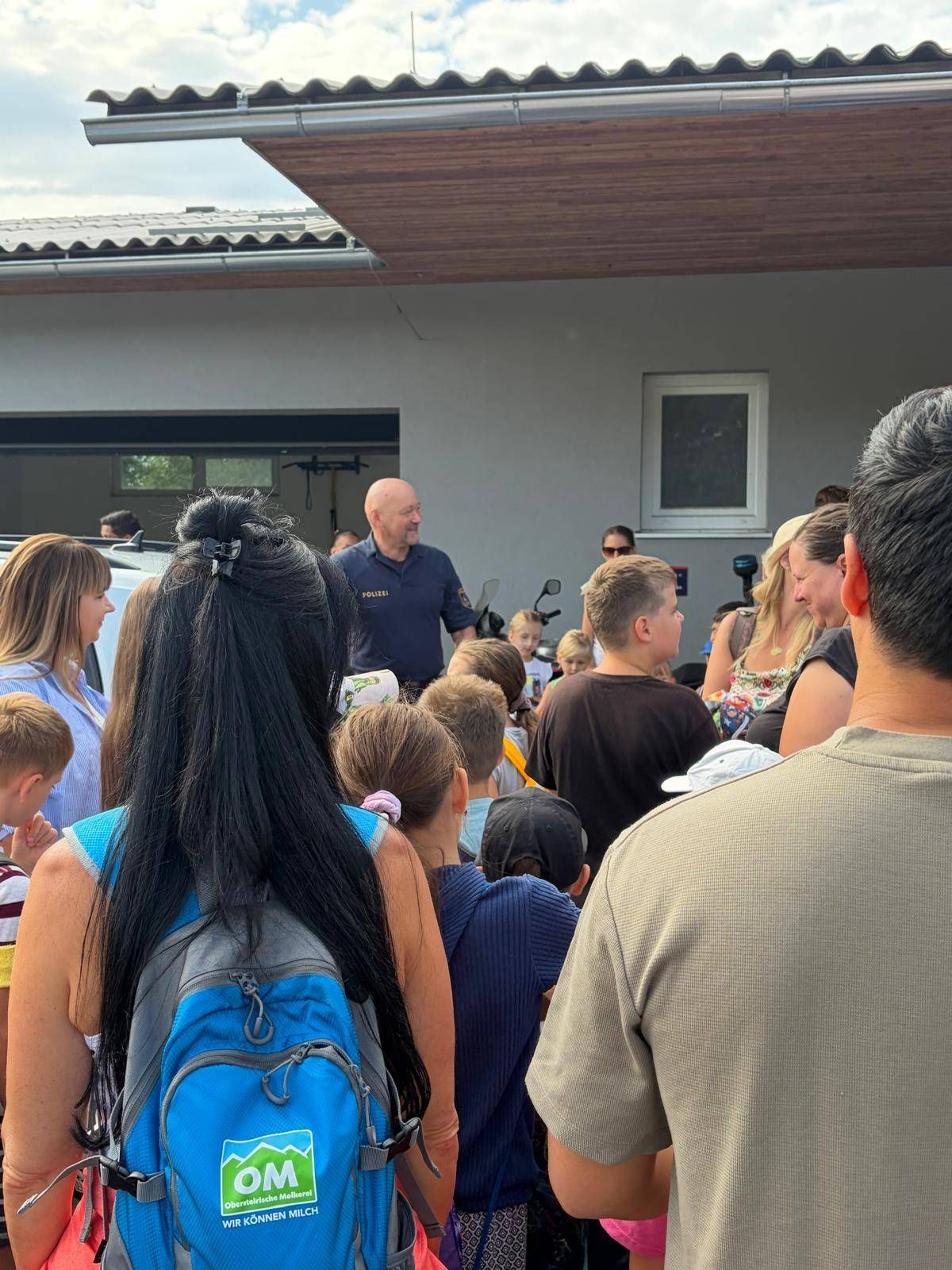 Eine Gruppe von Menschen, darunter Kinder, versammelt sich um einen Mann in blauer Uniform, möglicherweise ein Polizeibeamter, vor einem Gebäude.