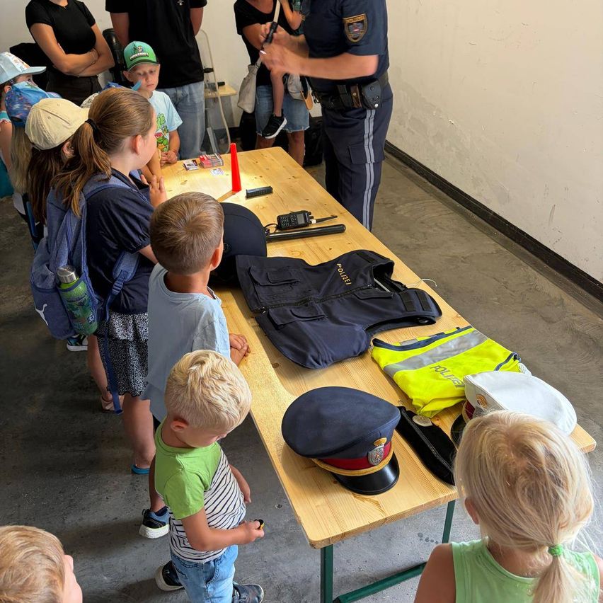 Eine Gruppe von Kindern und Erwachsenen versammelt sich um einen Tisch mit Polizeiuniformen und Ausrüstung. Ein Polizist erklärt den Kindern etwas.