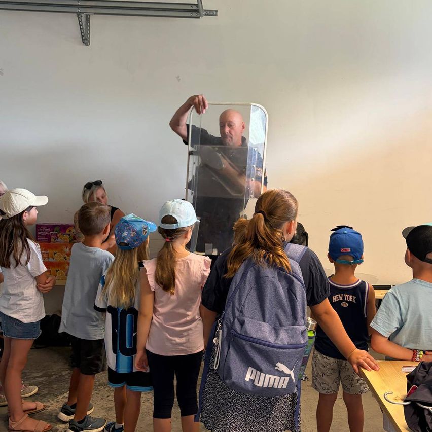 Eine Gruppe von Kindern und Erwachsenen ist um einen Mann versammelt, der einen durchsichtigen Schild hält, möglicherweise in einem Klassenzimmer. Die Kinder tragen Hüte und Turnschuhe, und einige haben Rucksäcke dabei. Der Mann scheint ihnen etwas zu erklären.