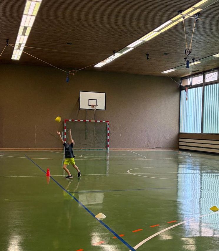 Ein junger Junge übt das Werfen eines gelben Balls in einer Sporthalle im Innenbereich, mit einem Basketballkorb im Hintergrund.