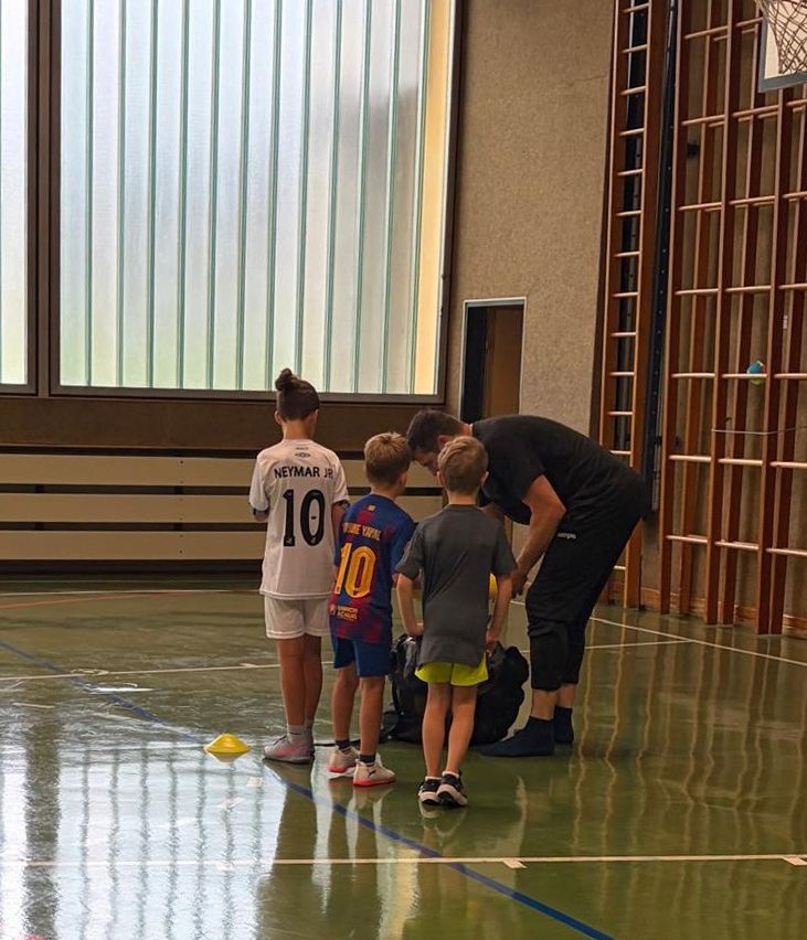 Eine Gruppe junger Jungen und ein Mann in einer Turnhalle, die Jungen tragen Sporttrikots und Turnschuhe, und der Mann hilft einem der Jungen bei seinen Schuhen.