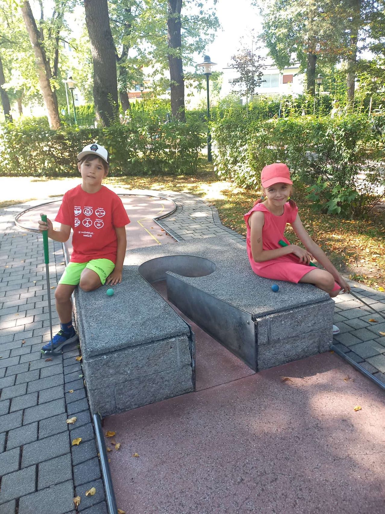 Zwei Kinder sitzen auf einer Betonbank im Park, halten Golfschläger in der Hand und lächeln für die Kamera.