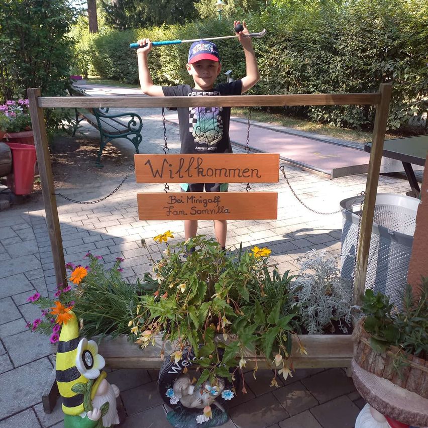 Ein junger Junge steht hinter einem Holzschild, auf dem 'Willkommen' steht, und hält einen Golfschläger in beiden Händen. Das Schild ist mit Ketten an einem Holzrahmen befestigt. In der Nähe befinden sich Topfpflanzen und ein Gartenzwerg.