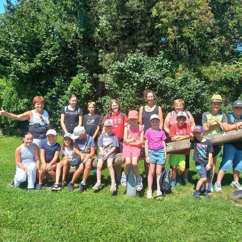 Eine Gruppe von Kindern und Erwachsenen posiert für ein Foto in einem Park, einige sitzen auf einem Baumstamm, andere stehen dahinter und alle lächeln.