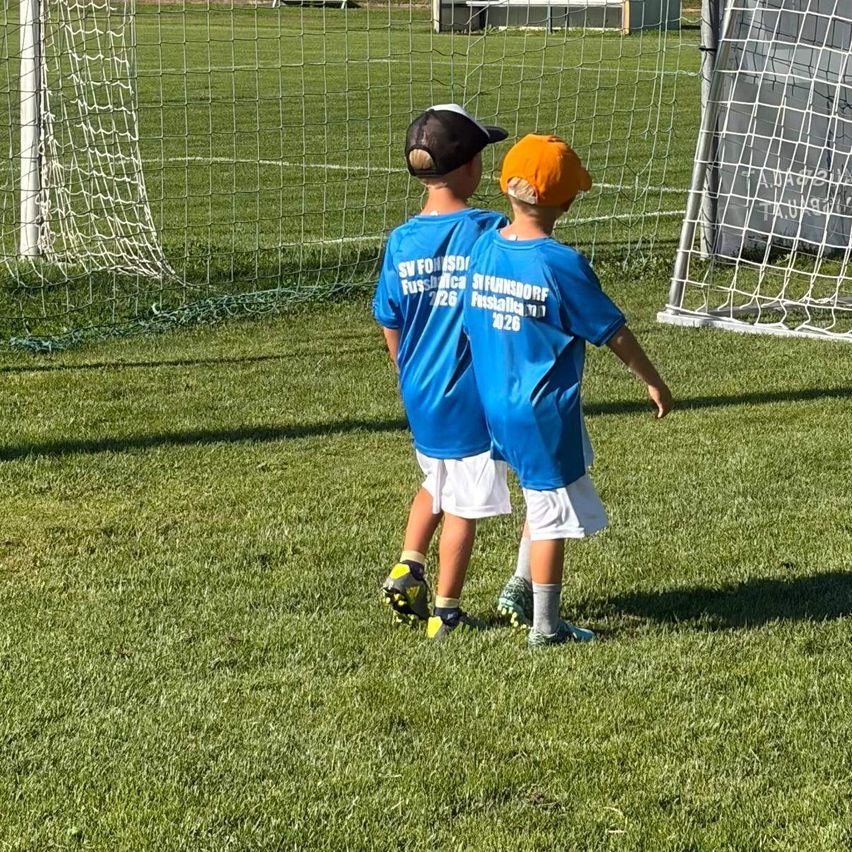 Zwei junge Jungen in blauen Fußballtrikots stehen an einem sonnigen Tag in der Nähe eines Tores.