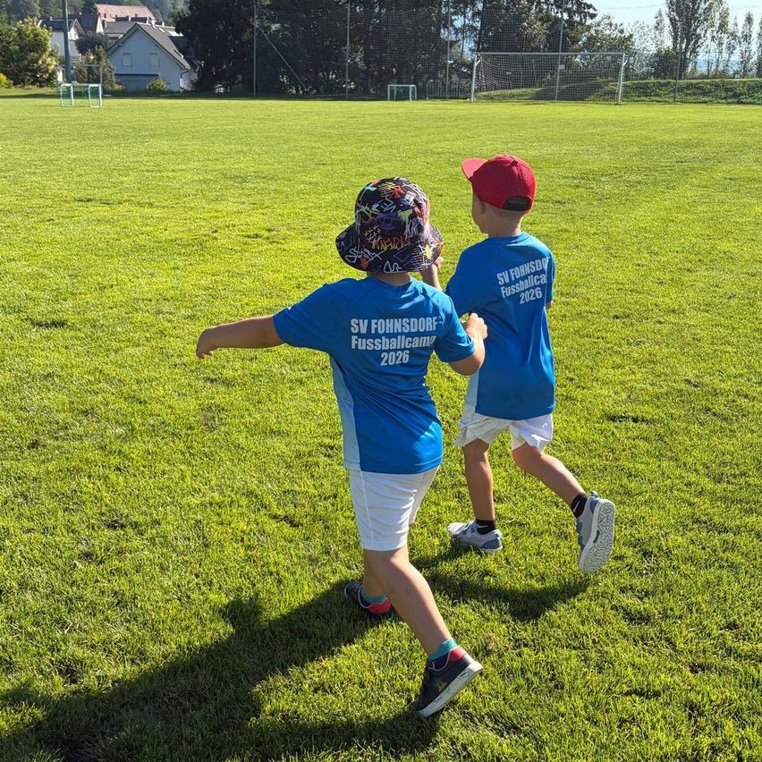 Zwei Jungen in blauen T-Shirts mit 'SV Fohnsdorf Fussballcamp 2026' darauf, tragen Mützen und Turnschuhe, rennen auf einem Rasenfeld mit einem Fußballtor im Hintergrund.