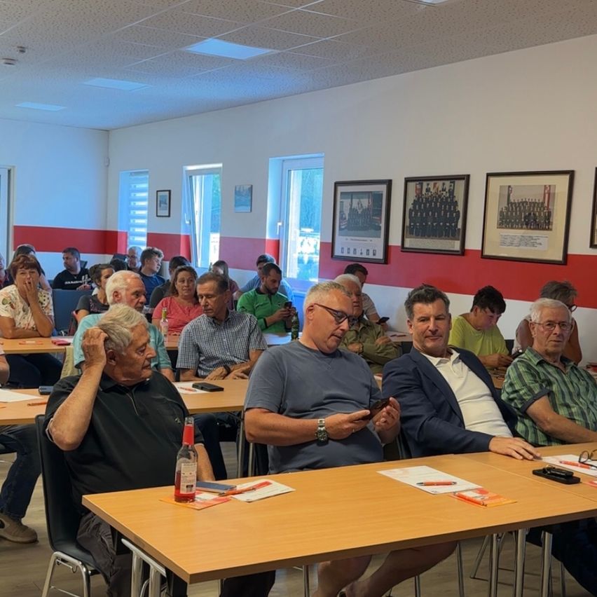 A group of people are seated around tables in a room. One person holds a phone. A few people are sitting on the chairs with their hands on their heads. A few frames are mounted on the wall.