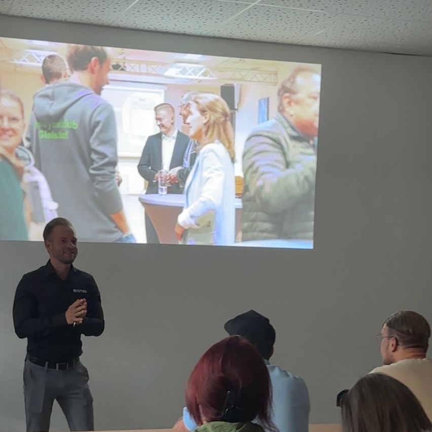 A man speaks in front of a projector screen showing people interacting, with others watching him.