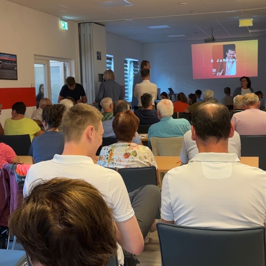 A group of people are seated in a room, watching a presentation on a projector screen. The room is well-lit, with tables and chairs arranged for the audience. A few people stand at the back.