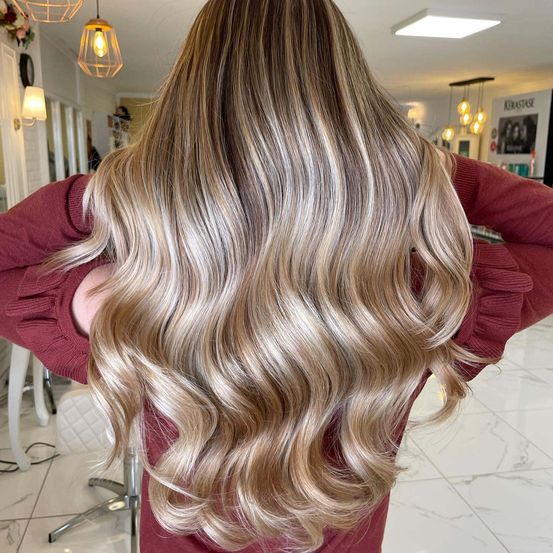 Eine Person mit langen, welligen blonden Haaren in einem Salon, trägt ein weinrotes Sweatshirt und schaut zurück. Der Salon hat Marmorböden, Pendelleuchten und ein Schild mit einem Bild.