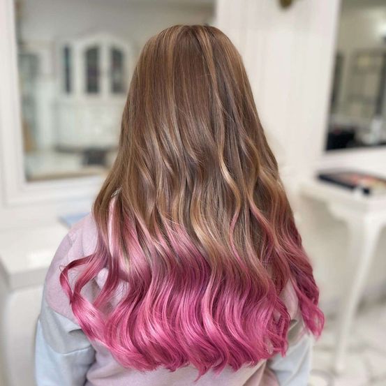 Eine Frau mit langen, gewellten Haaren, die in einem Farbverlauf von Pink zu Braun gefärbt sind. Sie trägt ein rosa Oberteil mit langen Ärmeln.