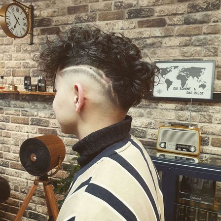 Ein Mann mit einem Mohawk-Haarschnitt steht in einem Barbershop, mit einer Uhr und einer Karte an der Wand hinter ihm.