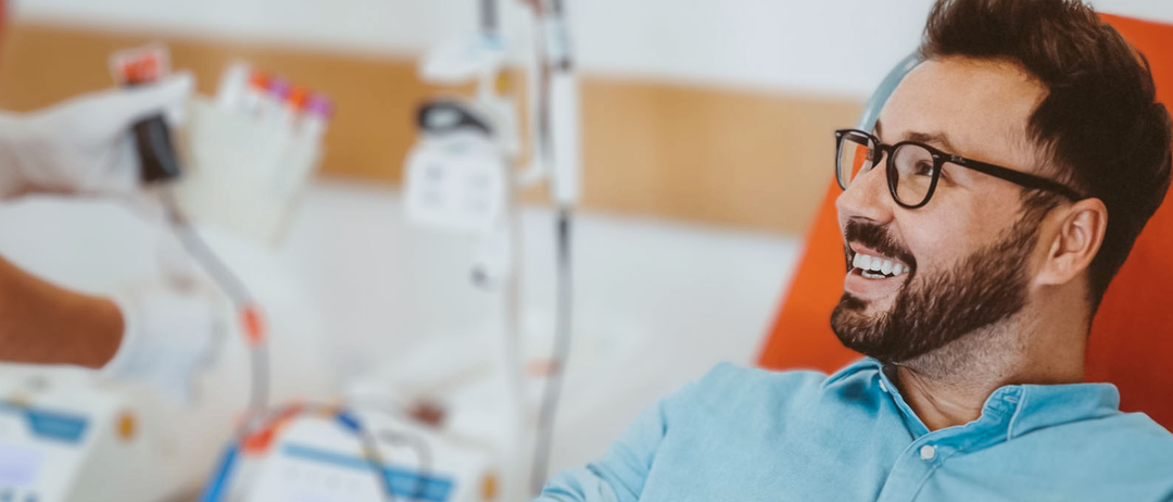 Ein Mann in einem blauen Hemd sitzt mit einem Verband am Arm in einem Krankenhausstuhl, lächelt und schaut auf sein Handy. Hinter ihm befinden sich medizinische Geräte und eine Krankenschwester in weißen Handschuhen. Der Text auf dem Bild lautet 'MI, 3.12.2025 17-20 Uhr Blutspenden in der vs Lannach'.