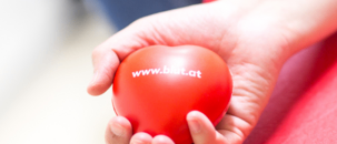 Eine Person hält einen roten Massageball mit der Aufschrift 'www.bat.at'. Die Hand liegt auf einer roten Oberfläche, und die Person scheint sich in einer Therapiesitzung zu befinden.