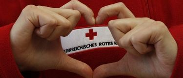 Eine Person bildet mit ihren Haenden eine Herzform. Sie traegt einen roten Pullover. Ein rotes Kreuz-Patch ist an ihrem Pullover befestigt.