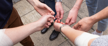 Eine Gruppe von Menschen in lässiger Kleidung mit verschiedenen Verletzungen steht eng beieinander, möglicherweise um einen Moment der Solidarität oder Unterstützung zu teilen, auf einem Betonboden.