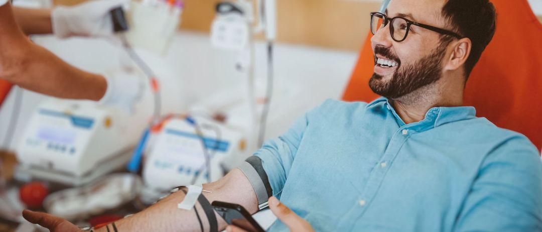 Ein Mann sitzt in einem Krankenhausstuhl, trägt eine Brille und ein blaues Hemd. Er lächelt und hält ein Telefon. Hinter ihm sind medizinische Geräte zu sehen, und ein Verband befindet sich auf seinem Arm.
