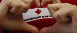 Eine Person in einem roten Pullover formt mit den Händen ein Herz, das ein weißes und rotes Emblem mit einem roten Kreuz in der Mitte zeigt. Der Text lautet 'INTERREICHISCHES ROTES K'.