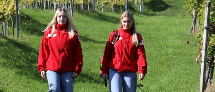 Vier Personen, die rote Jacken und Jeans tragen, stehen auf einem Rasen. Zwei Frauen stehen und lächeln, während ein Mann auf dem Boden sitzt. Eine weitere Frau liegt mit einem Lächeln auf dem Boden.