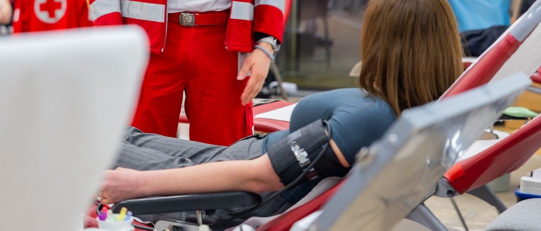 Ein Mann in rotem und weißem Uniform steht neben einer sitzenden Person mit einem Blutdruckmessgerät am Arm. Dahinter steht eine weitere Person. Der Bereich ist gut beleuchtet.