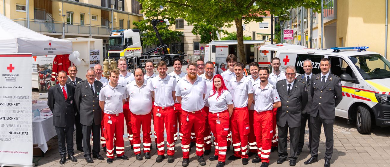 Eine Gruppe von Rettungskräften in roten Uniformen steht auf einem Stadtplatz, mit mehreren Fahrzeugen und Gebäuden im Hintergrund.